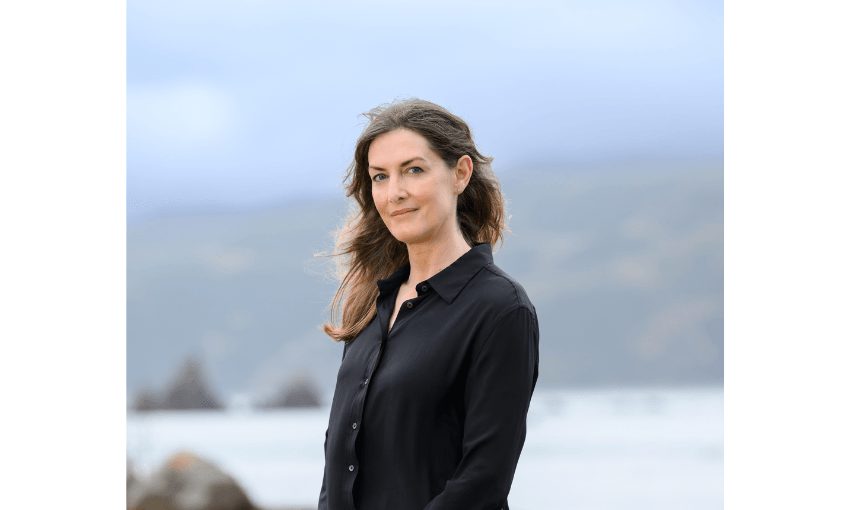 Photograph of a woman with long brown hair standing outside near a beach. She is looking at the camera.